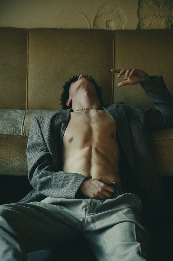A dramatic portrait of a shirtless man in a gray blazer lounging on a sofa.